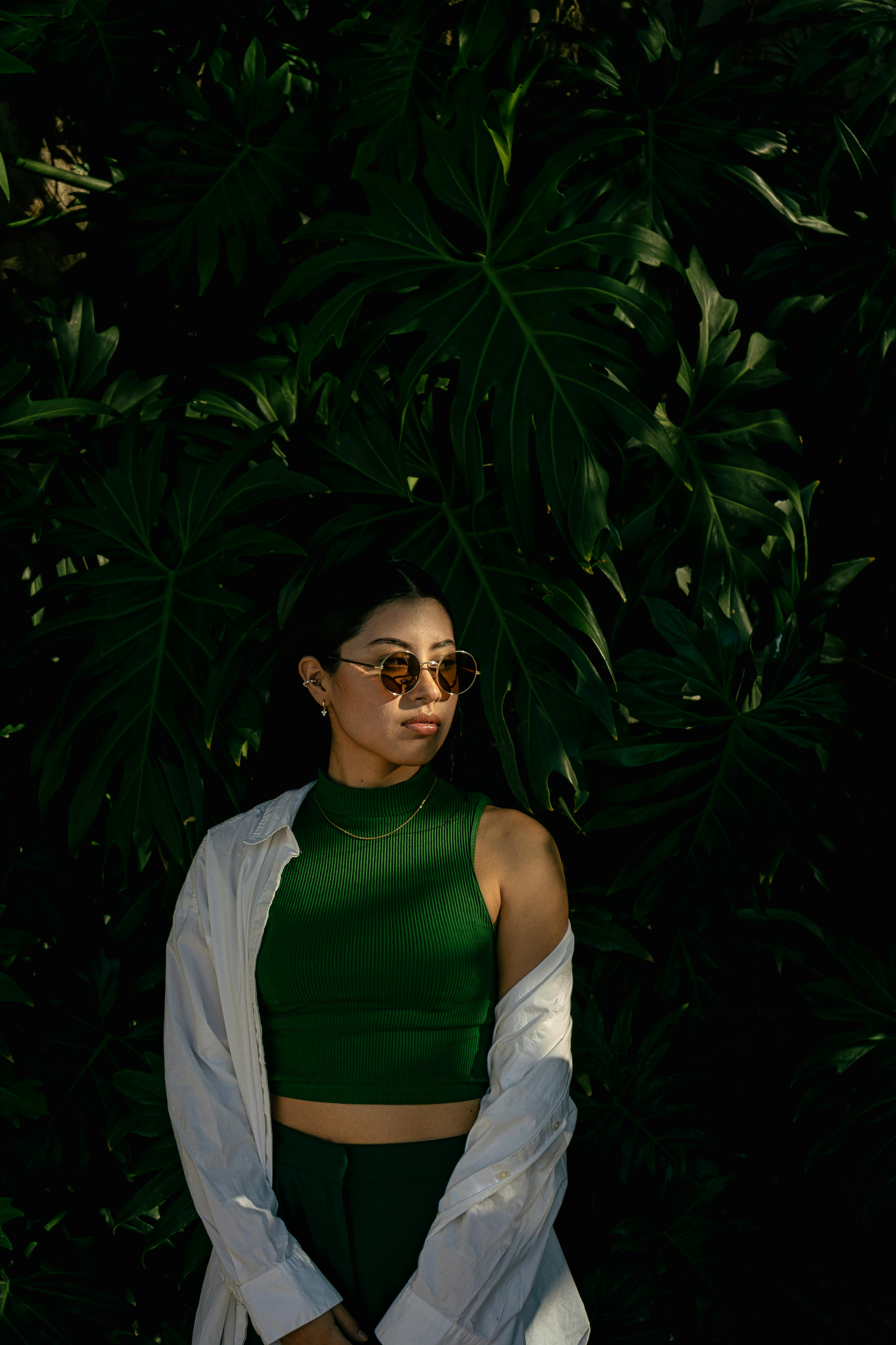 Fashionable woman poses elegantly in sunglasses and green outfit against lush leaves.