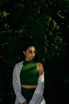 Fashionable woman poses elegantly in sunglasses and green outfit against lush leaves.