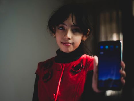 A young girl in a red dress shows a smartphone indoors, smiling softly.