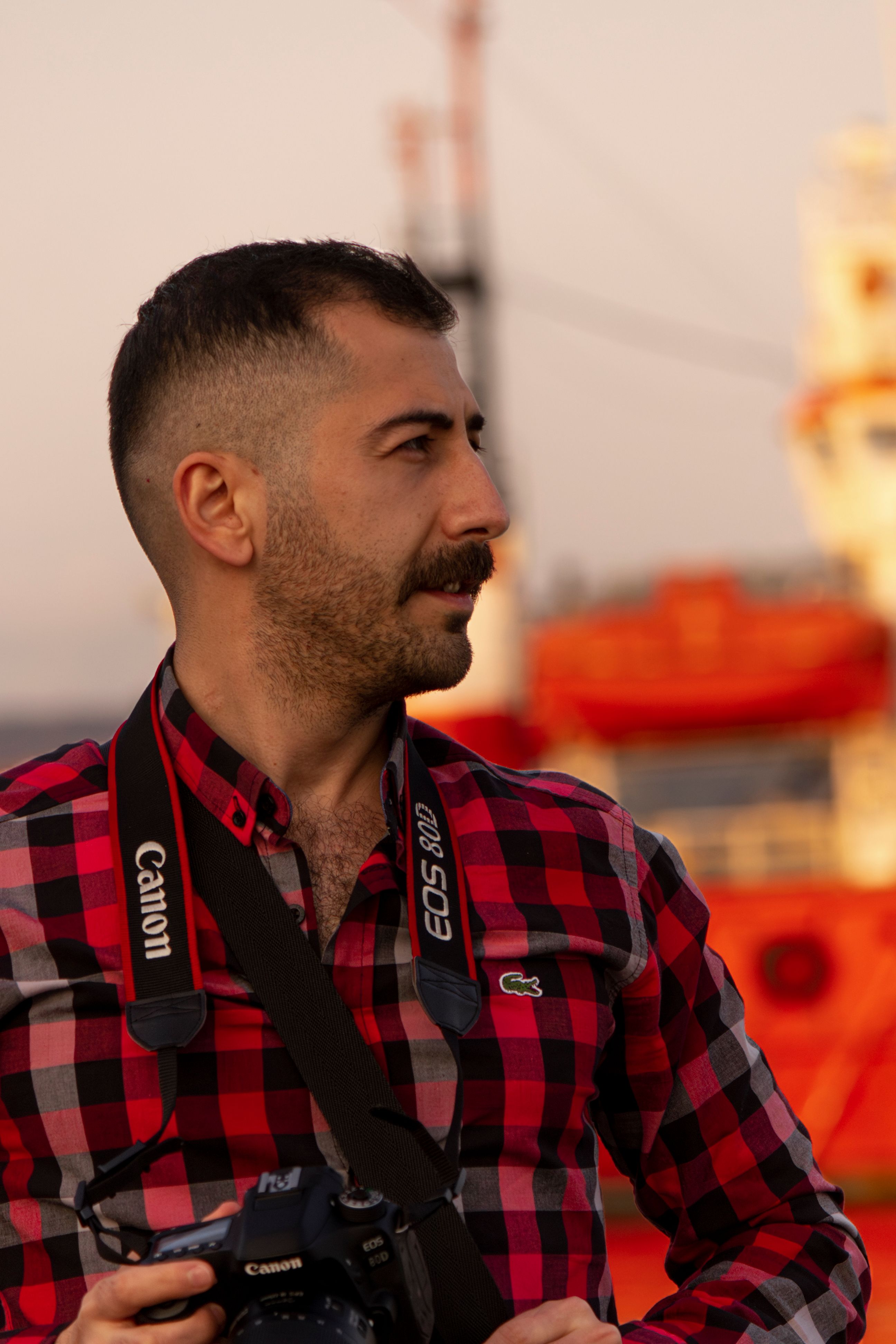 Male photographer with Canon camera standing outdoors with a vibrant orange ship in the background at sunset.