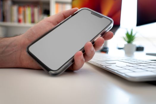 Close-up of a hand holding a smartphone with blank screen over a desk with keyboard and plant.