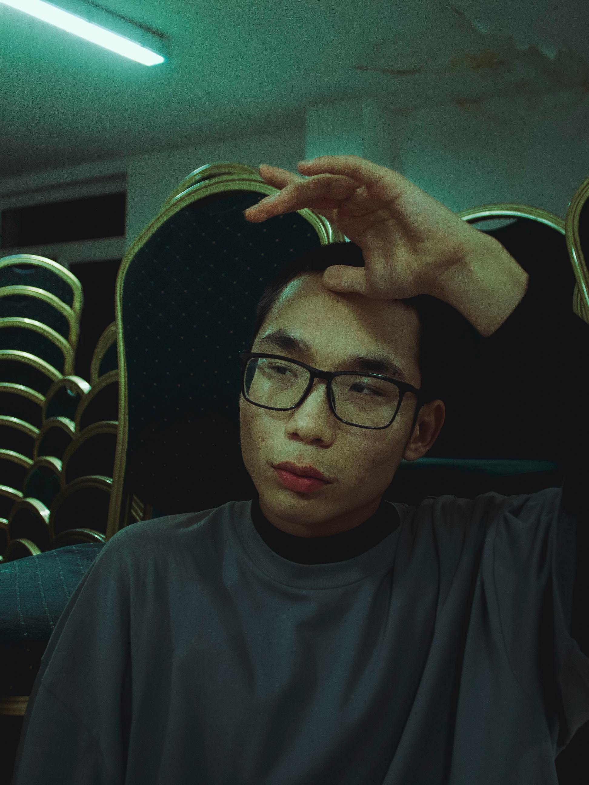 A man with eyeglasses looks thoughtful in a dimly lit room, seated indoors.