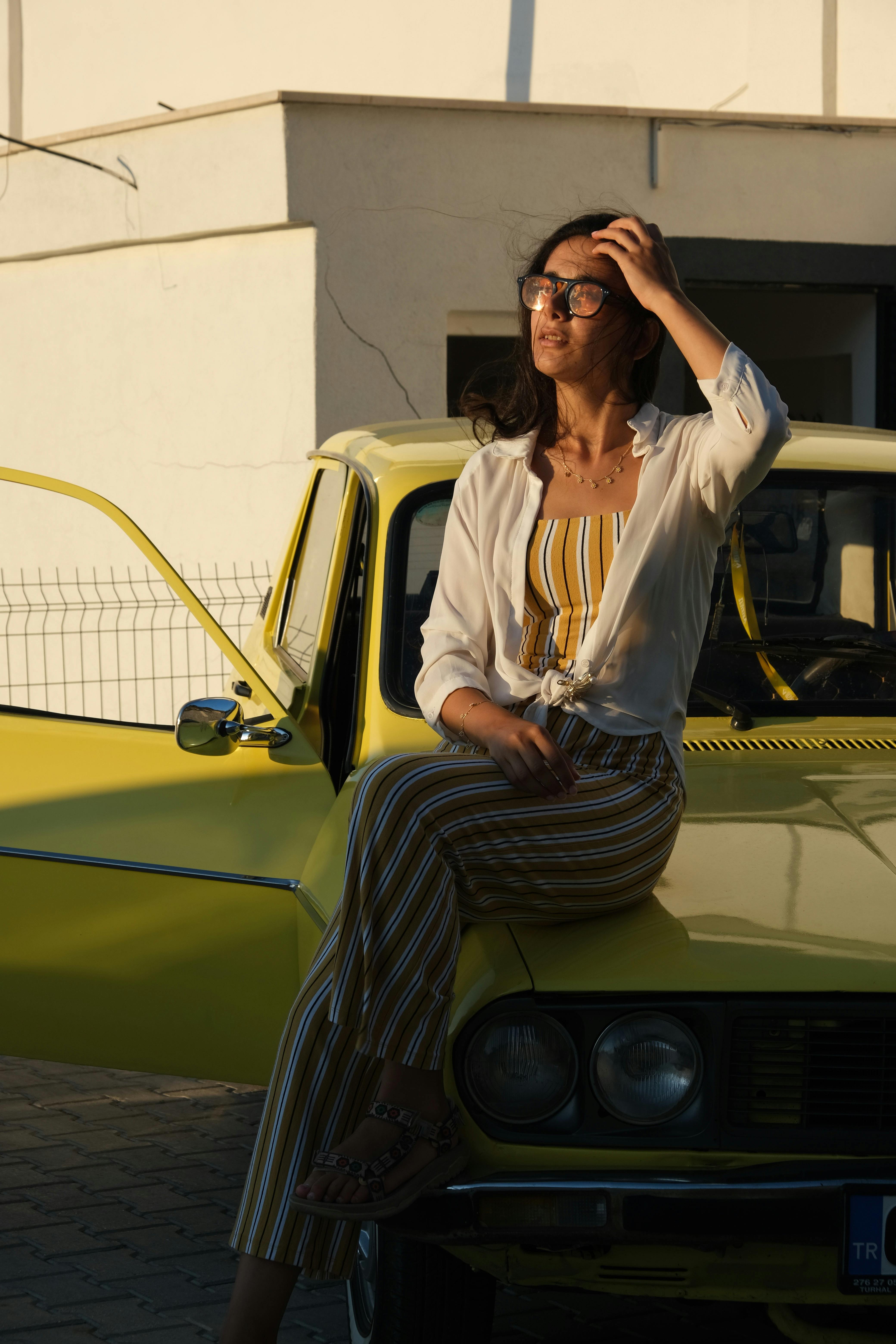 A fashionable woman sits on a yellow vintage car at sunset, wearing a striped outfit.