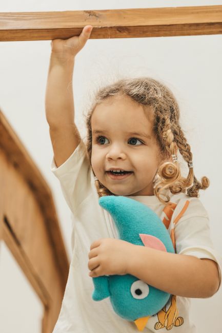 Happy child with braids holding a stuffed penguin toy indoors, arm raised playfully.