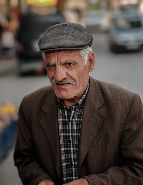 Free Close-up portrait of an elderly man with mustache wearing a flat cap and jacket, standing on a street. Stock Photo