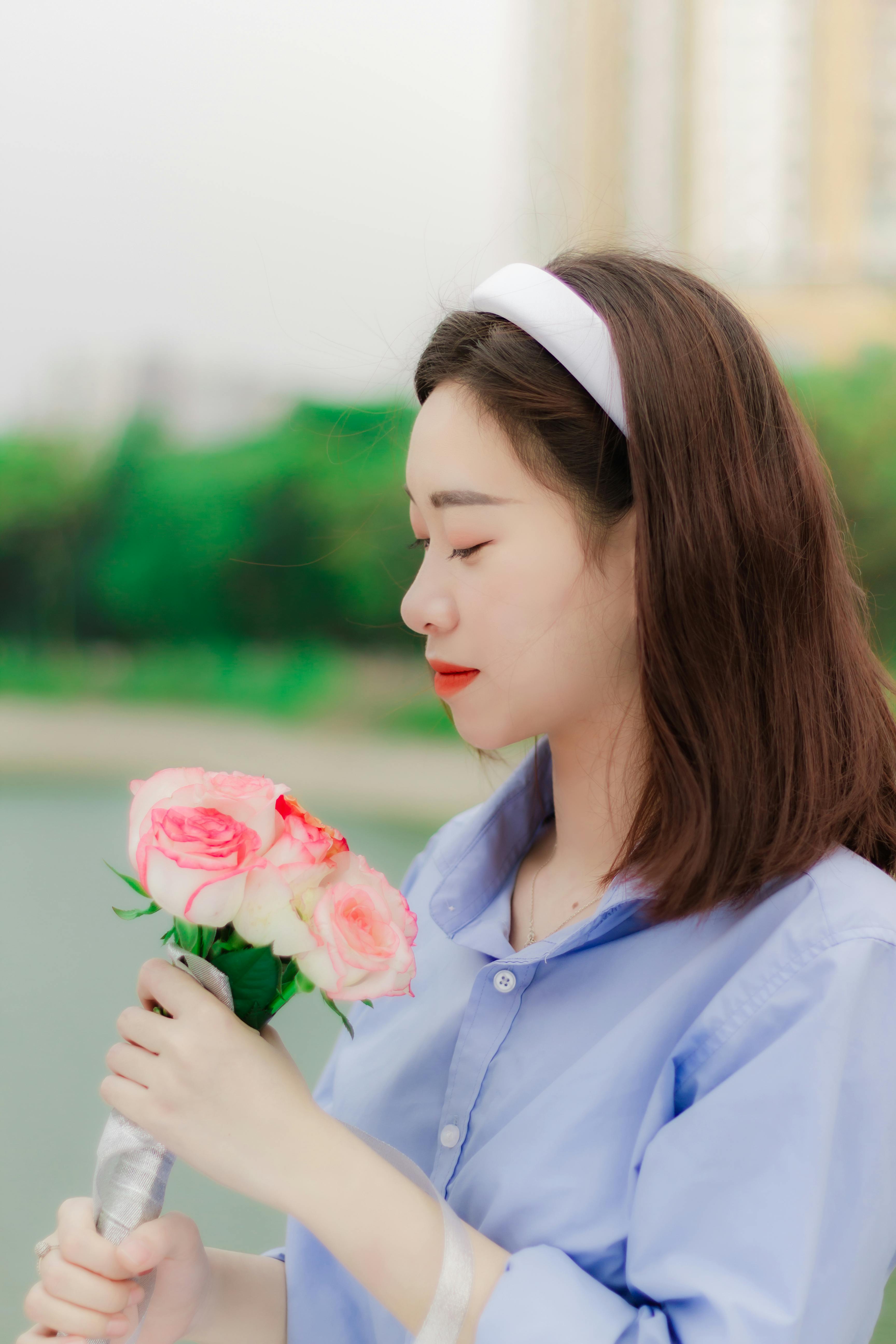 Young woman in blue shirt holding pink roses, standing outdoors with serene expression.