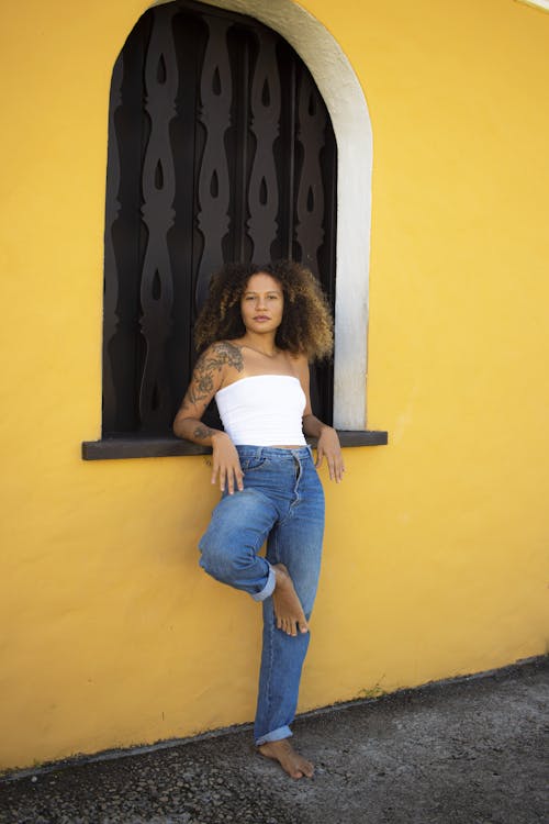 Young Woman in a White Top and Jeans Standing Barefoot in front of a Yellow Wall 