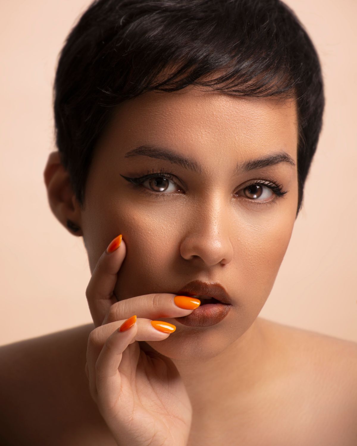Studio portrait of a woman with striking makeup and orange nails against a beige background.