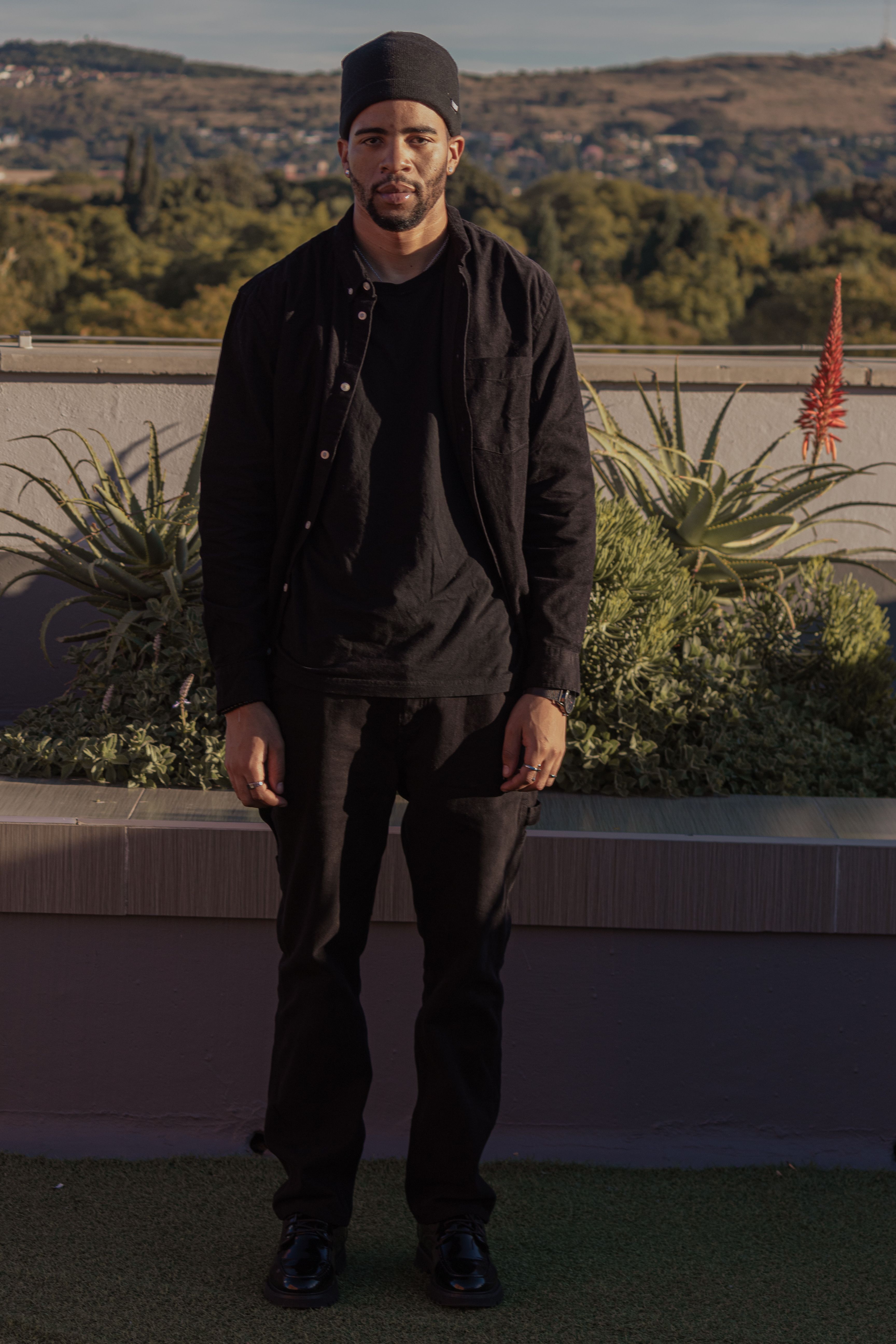 A man with a mustache stands in front of plants on a rooftop, outdoor fashion shot.