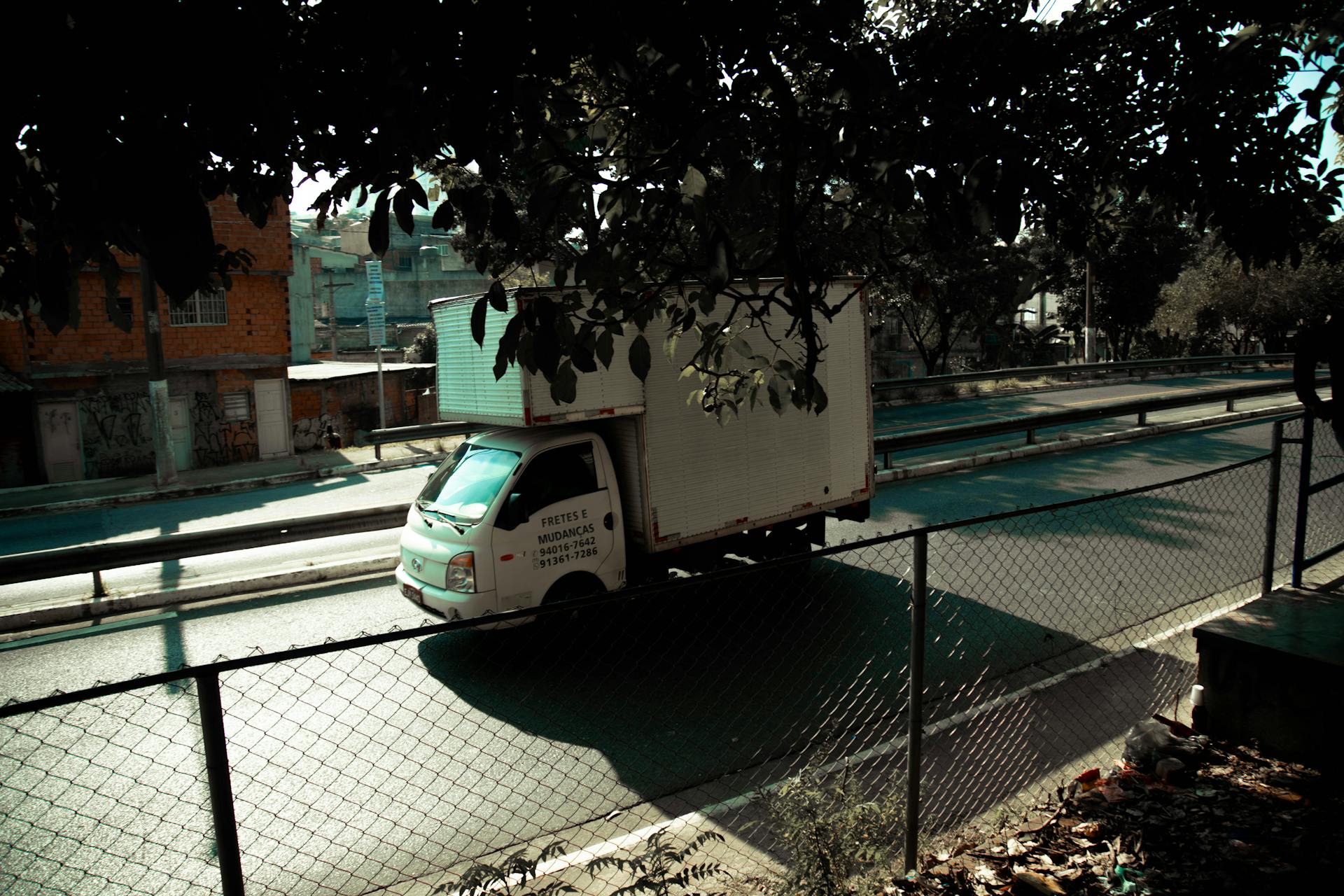 Delivery truck on sunlit urban road, framed by fence and trees, showcasing urban life contrast.