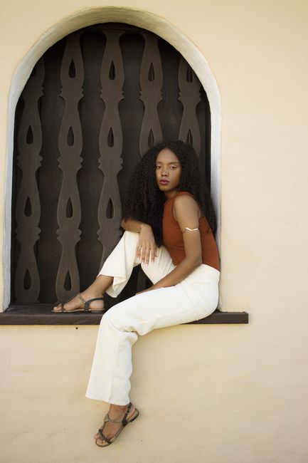 Elegant portrait of a woman in white pants sitting by an ornate window in Porto Seguro, Brazil.
