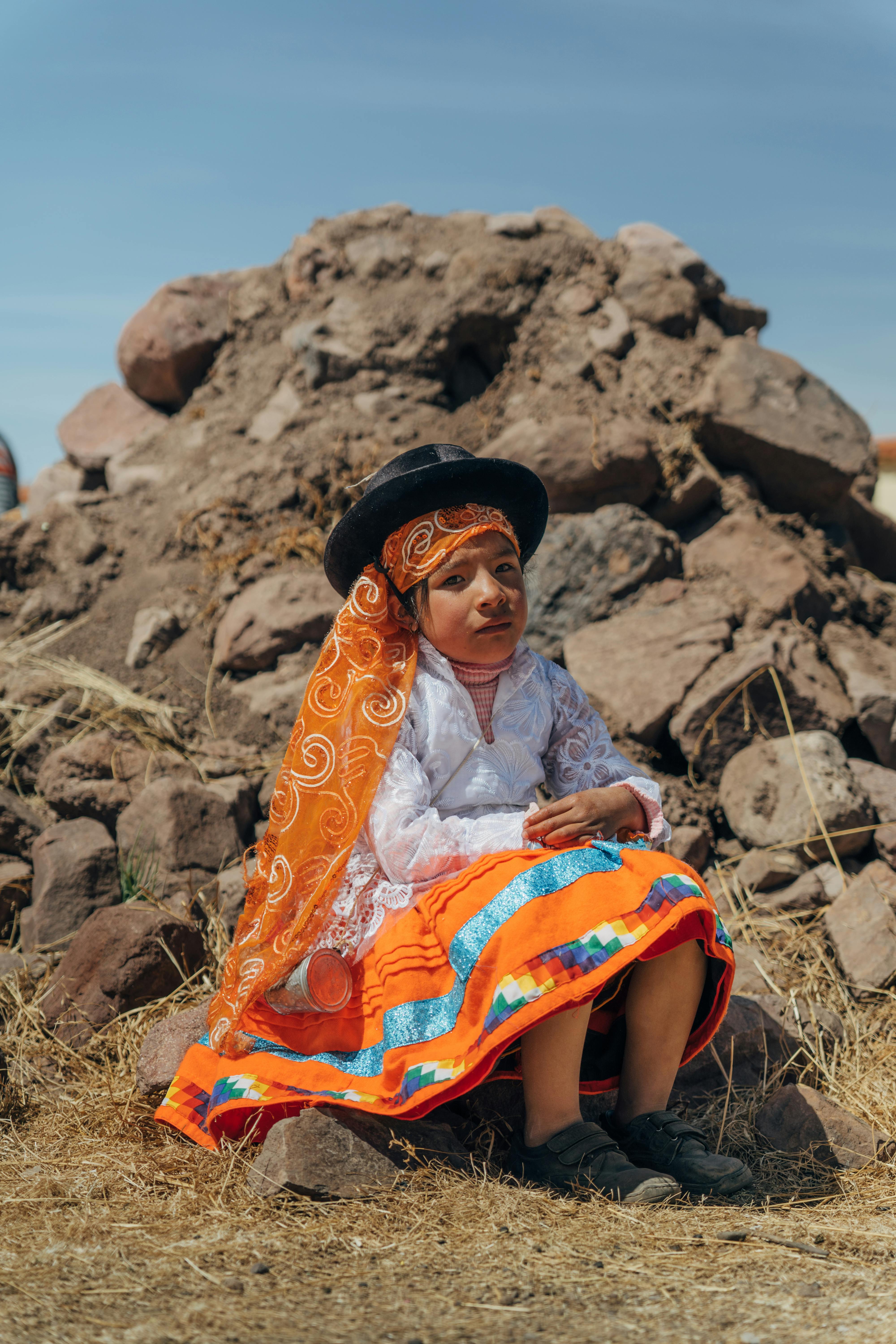 A young girl in vibrant traditional clothing sits outdoors against a rocky backdrop in Puno, Peru.