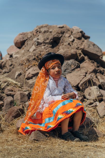 A young girl in vibrant traditional clothing sits outdoors against a rocky backdrop in Puno, Peru.