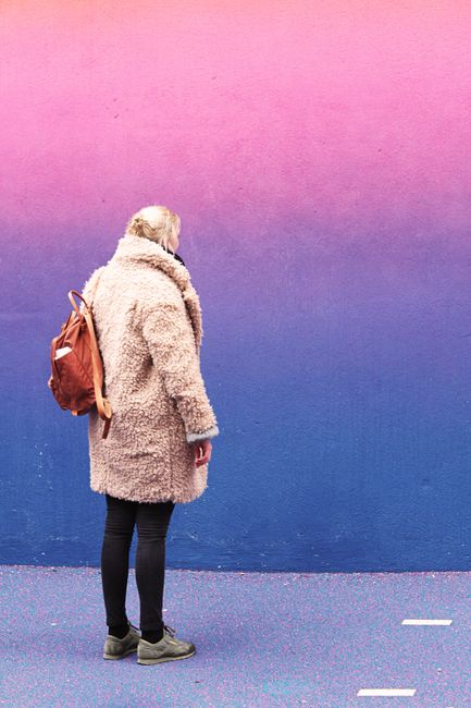 Woman in stylish fur coat and backpack stands by vibrant pink and blue wall.