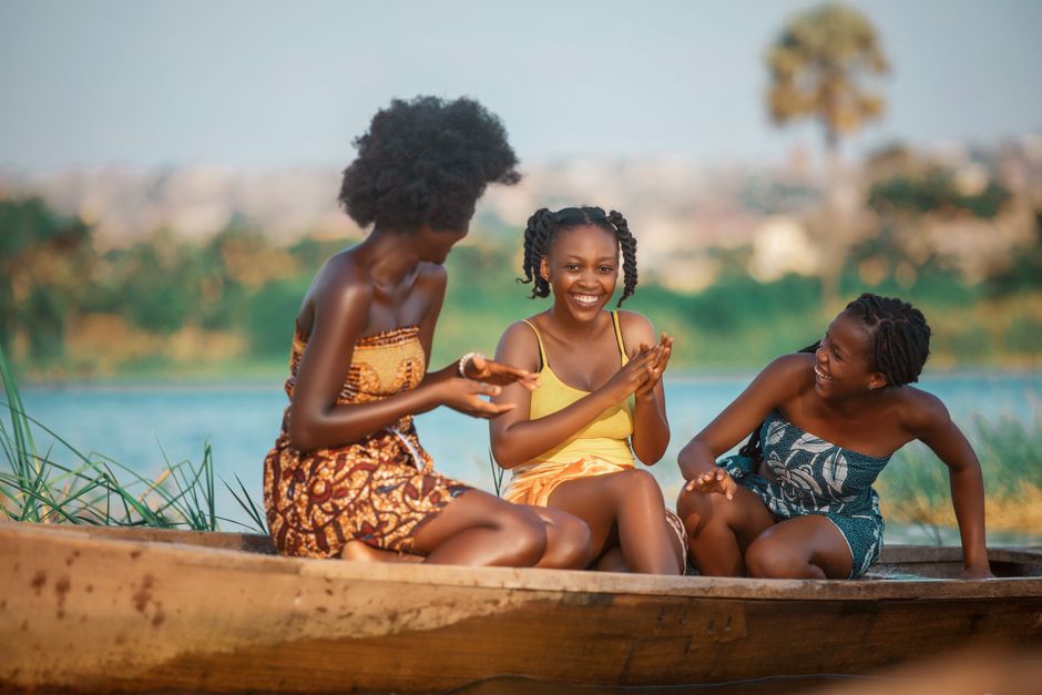 Three young women laughing and sitting in a canoe by the river in Accra, Ghana, enjoying a sunny day.