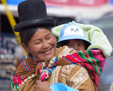 A joyful Bolivian woman in traditional clothing carrying a child in a vibrant shawl, outdoors.