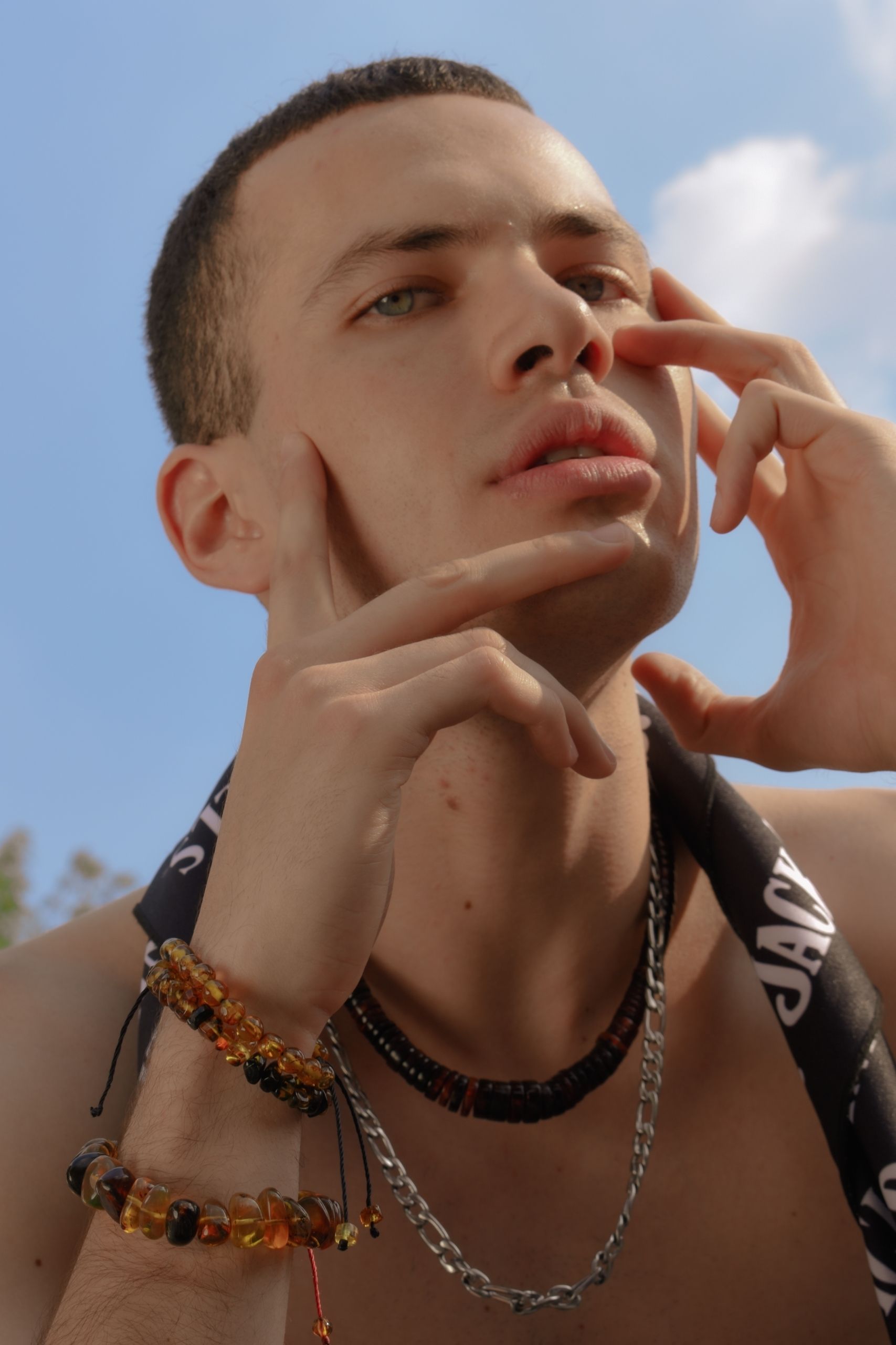 A shirtless man outdoors in Ciudad de México, wearing bracelets and a necklace, looking contemplative.