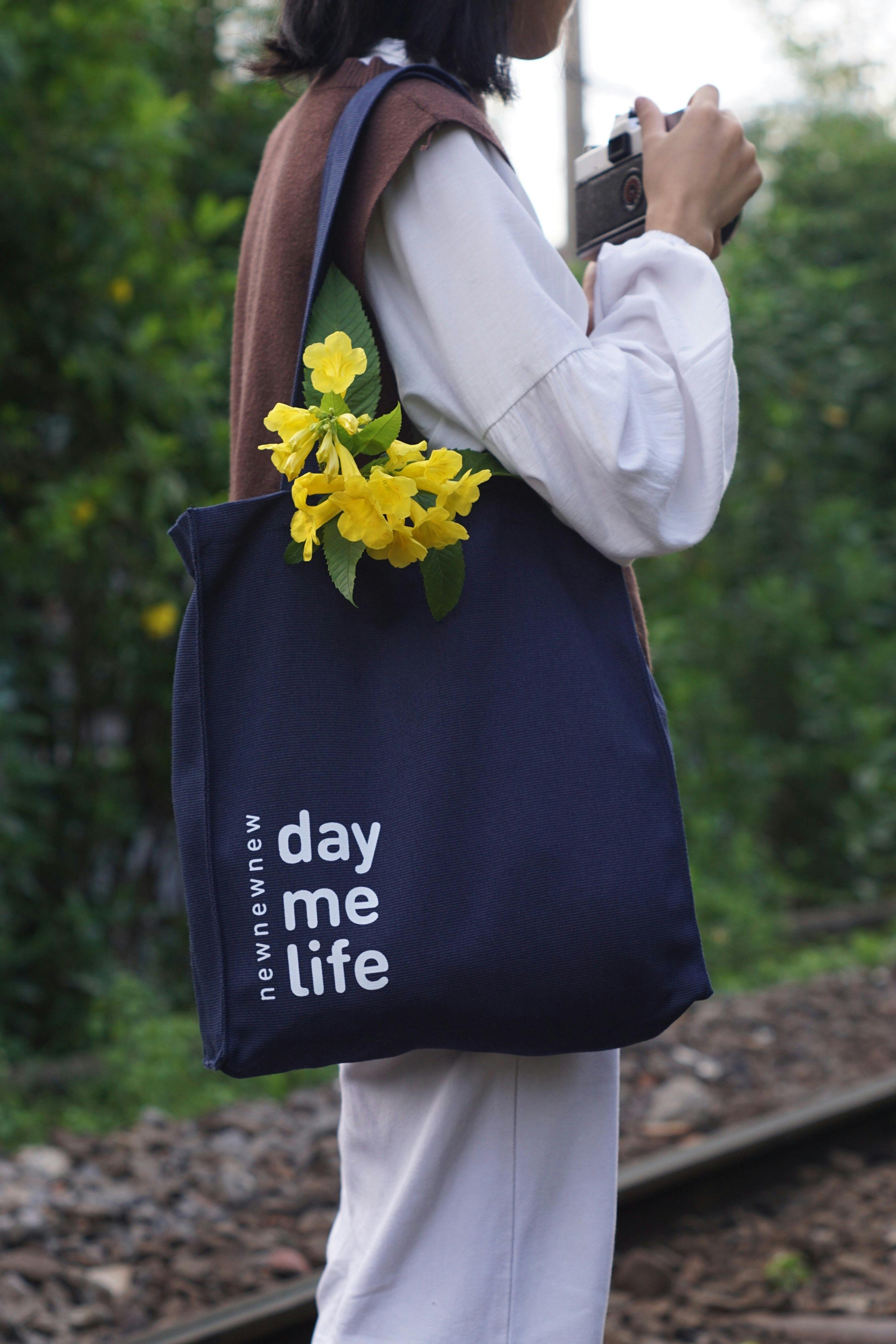 Woman standing by railway with a navy tote bag and yellow flowers.