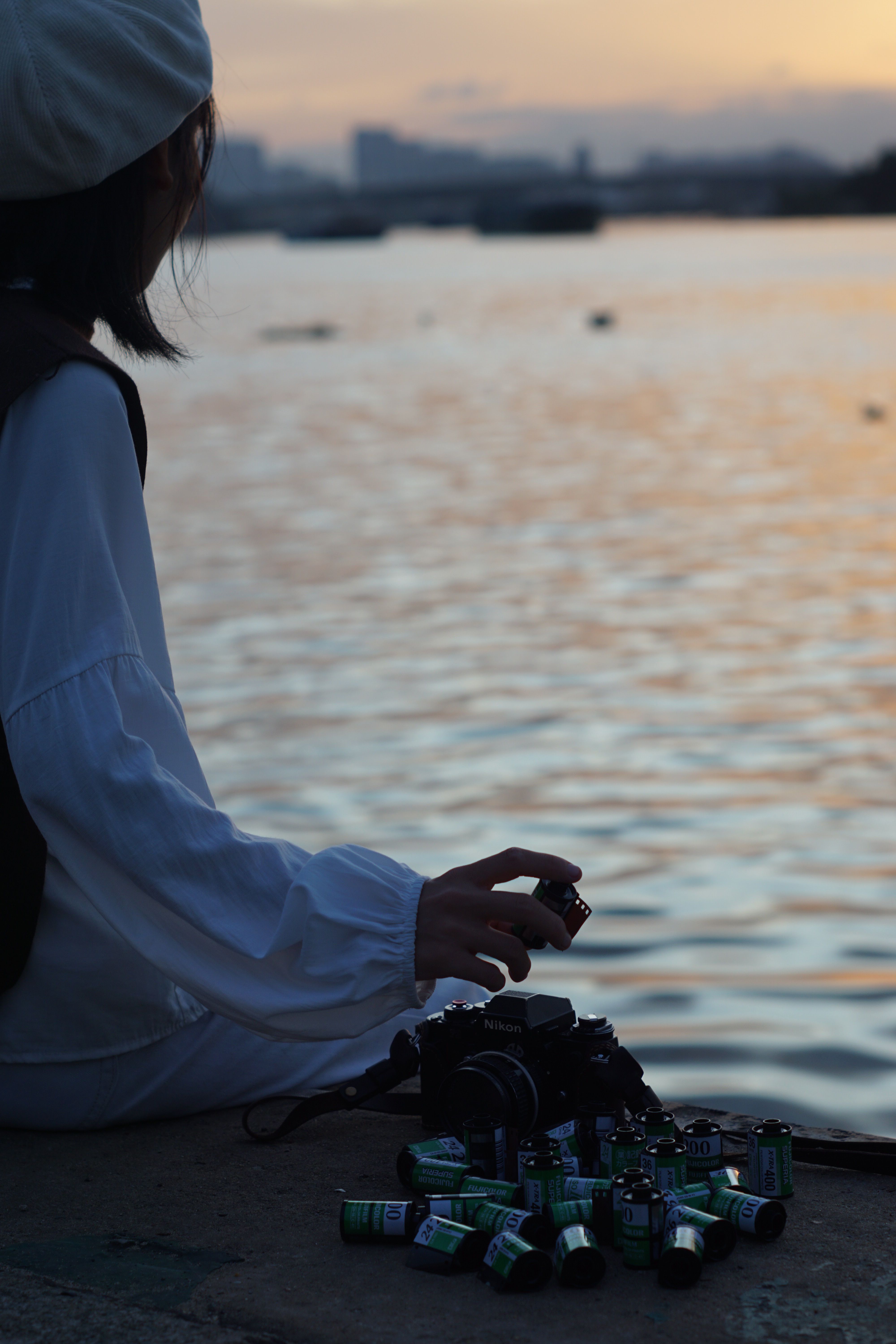 A photographer sits by the water at sunset, surrounded by film canisters, creating a serene and nostalgic scene.