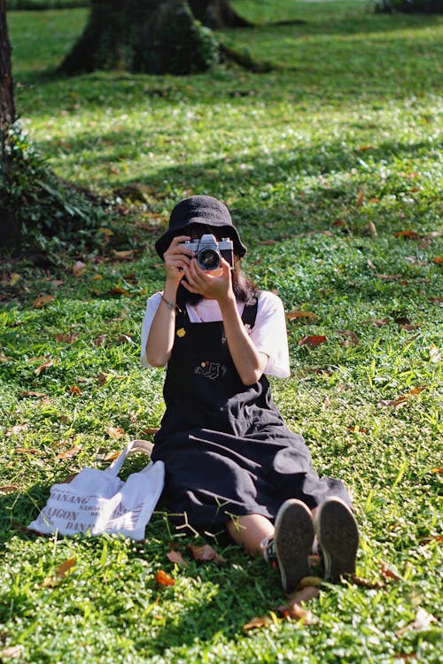 Young Woman in Black Dress with Suspenders Sitting on a Lawn with a Photo Camera