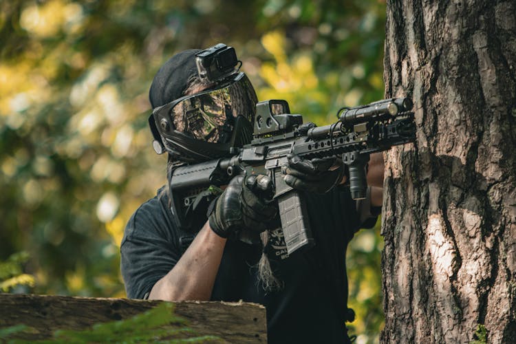 Man In Protective Paintball Mask Aiming A Weapon Hiding Behind A Tree