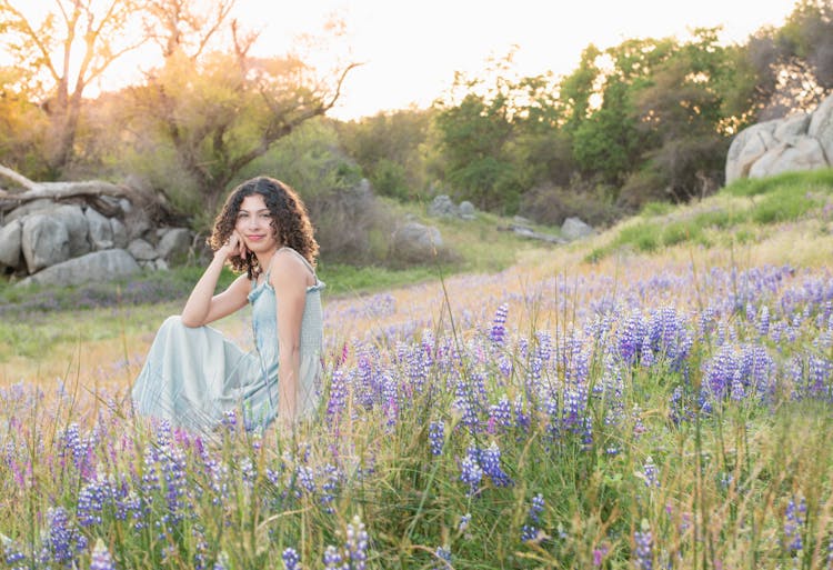 Young Woman In Pastel Blue Summer Dress Sitting In A Mountain Meadow With Blue Lupine Flowers