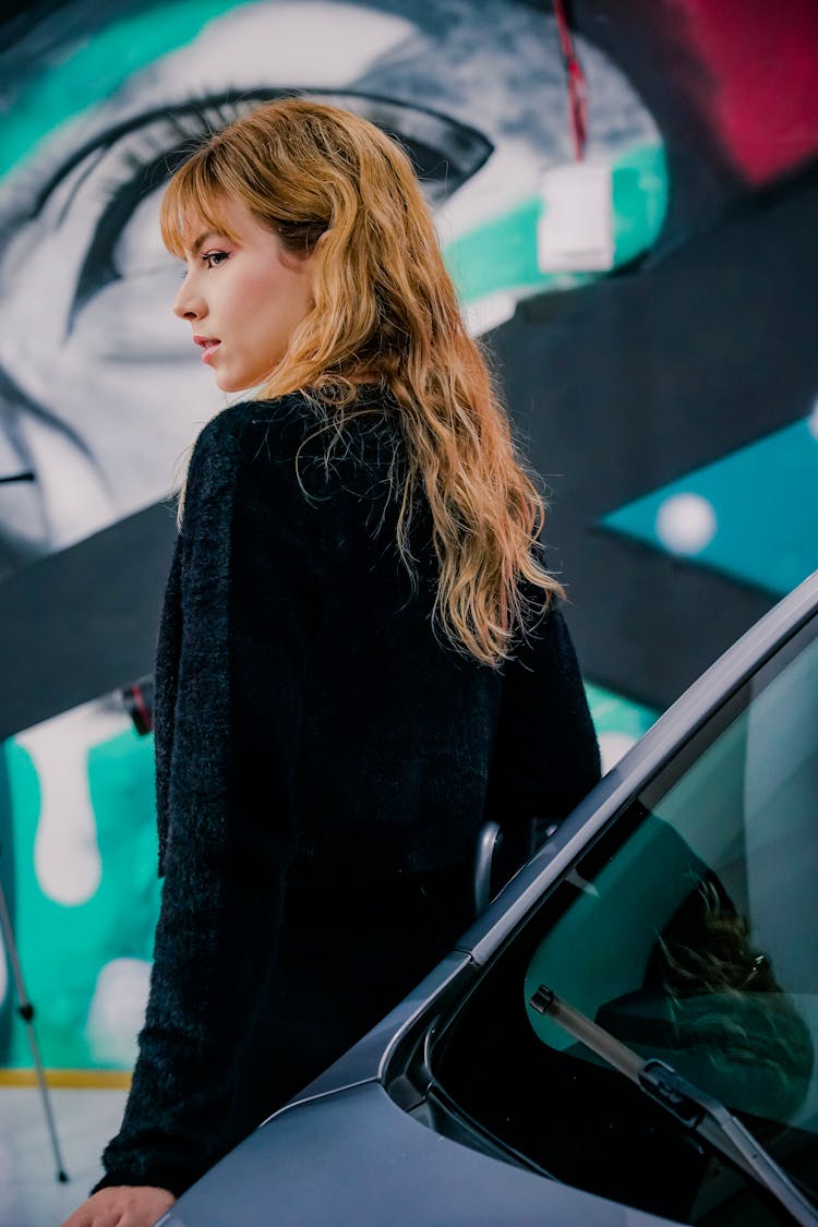 Young Woman Standing Next To A Car In A Garage With A Mural On The Wall 