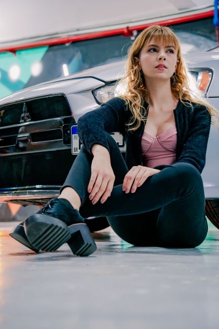 A young woman with blonde hair poses confidently on the garage floor in front of a white car.
