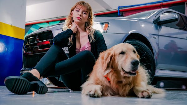 Young woman and golden retriever dog sitting beside a parked car in a garage setting.
