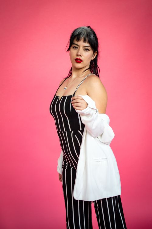 Free Fashionable woman in striped jumpsuit and white cardigan poses confidently on a pink backdrop. Stock Photo