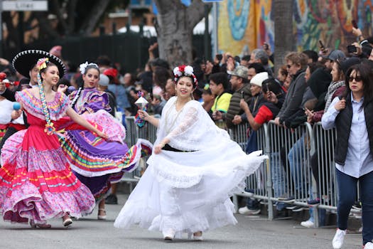 Women in colorful traditional attire dance in a lively street parade watched by a crowd.