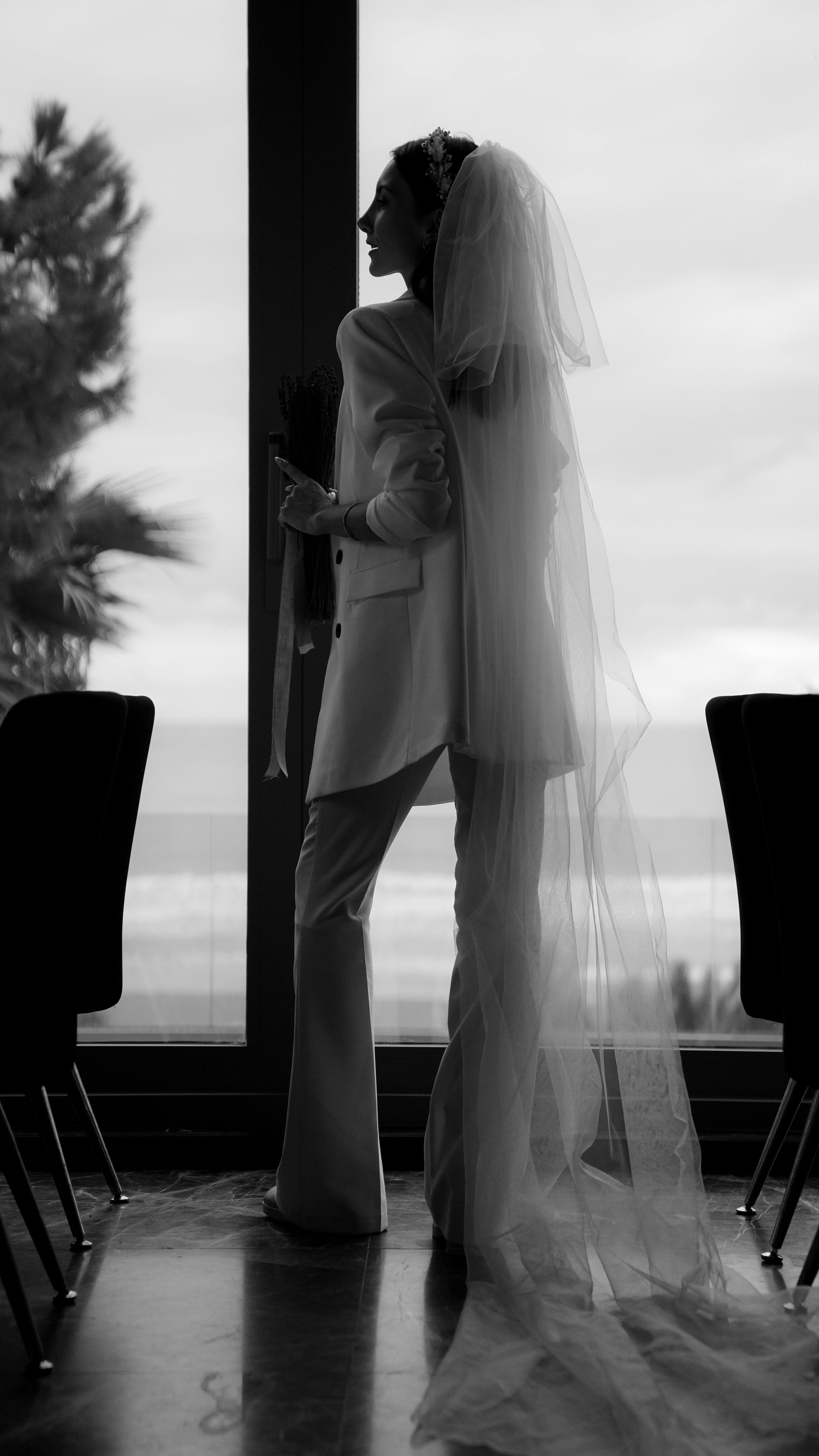 A bride in a tailored suit and veil stands in silhouette beside a window