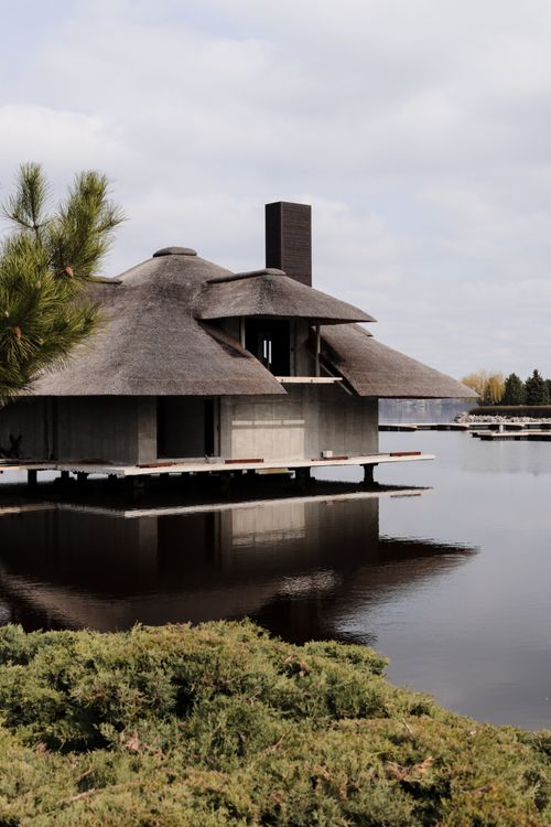 Free A peaceful lake house with a unique thatched roof reflected on still water, surrounded by nature. Stock Photo