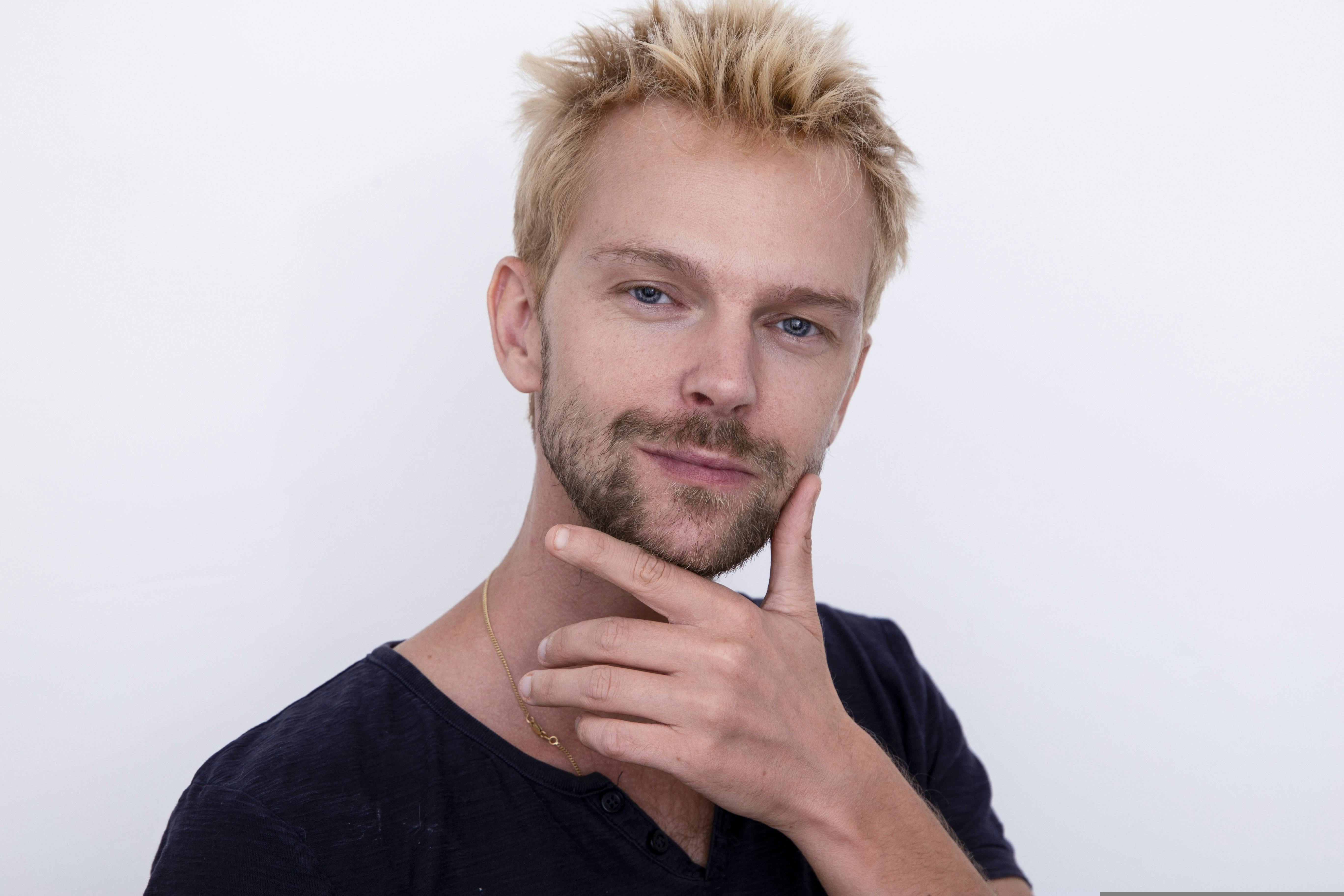 Portrait of a confident blonde man posing with hand on chin against a white background.