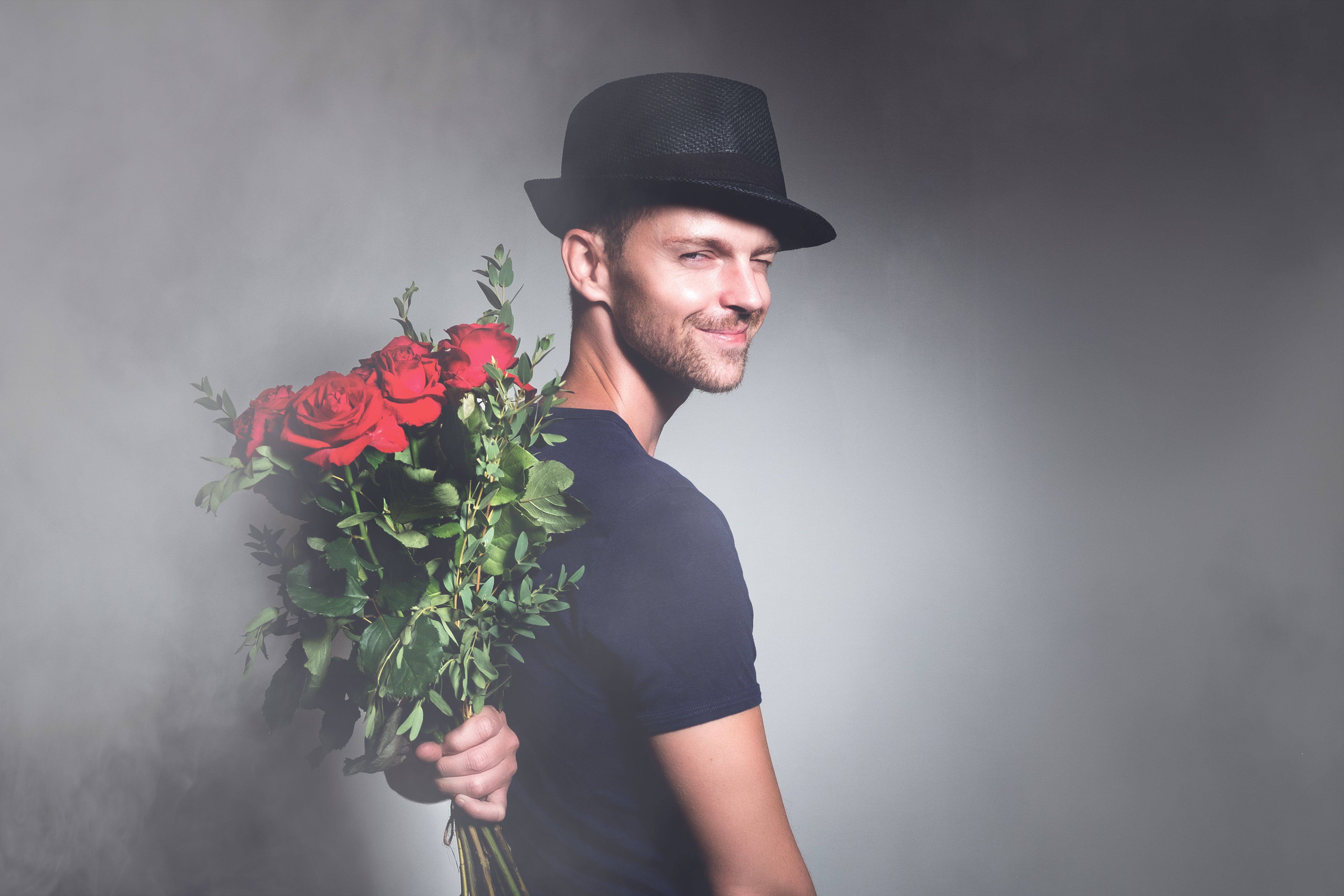 A stylish man in a hat holds a bouquet of red roses, exuding charm and elegance.