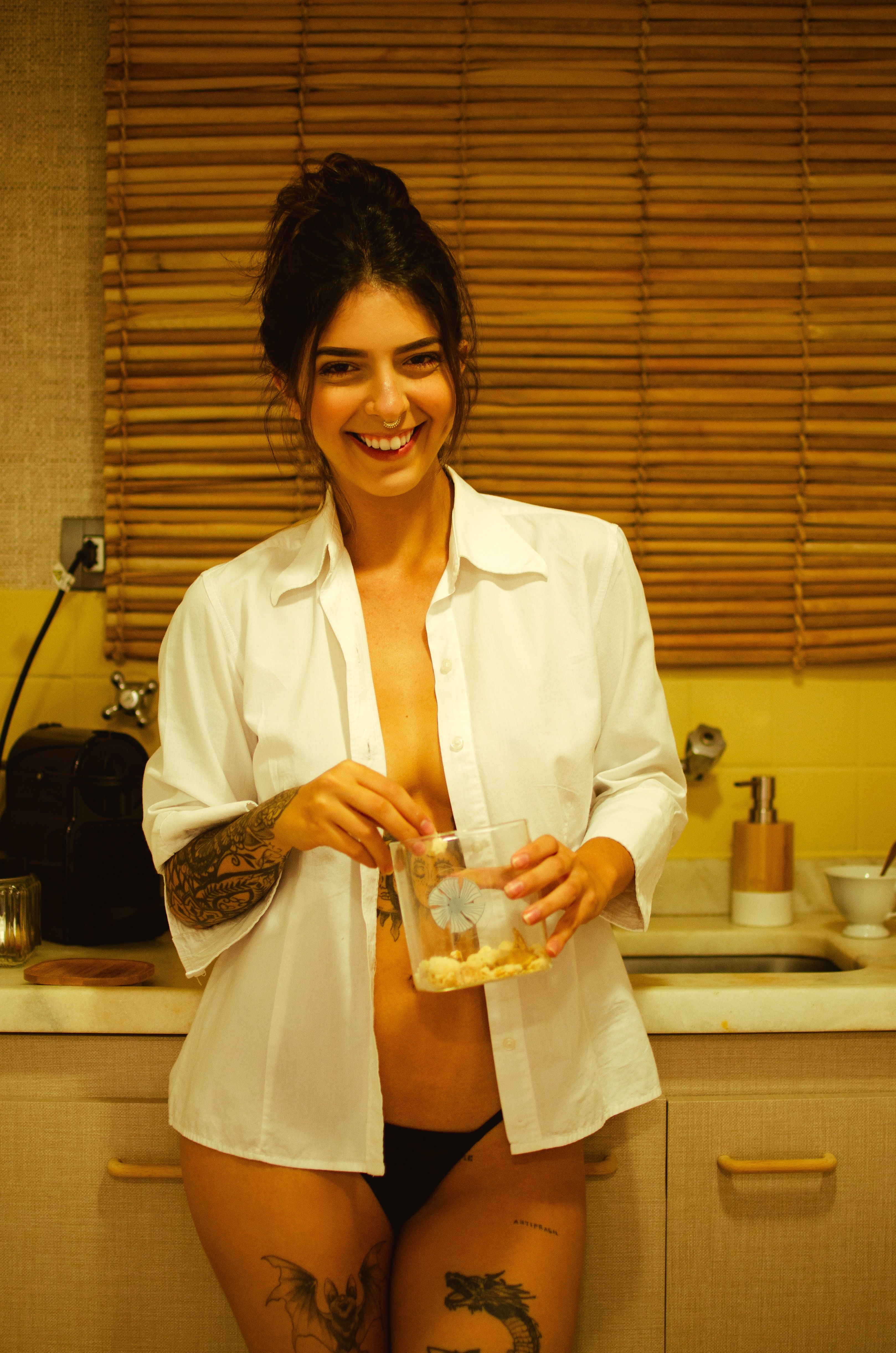 Smiling woman in lingerie and open shirt holding a glass of cereal in a cozy kitchen setting.