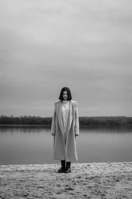 Artistic black and white portrait of a woman standing by a calm lake under an overcast sky.
