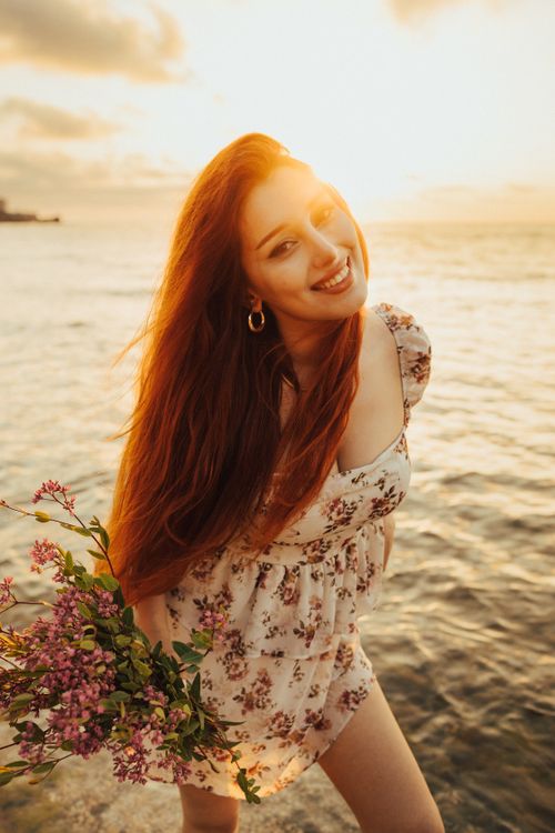 Free A joyful woman with red hair, holding flowers by the sea during a vibrant sunset in Türkiye. Stock Photo