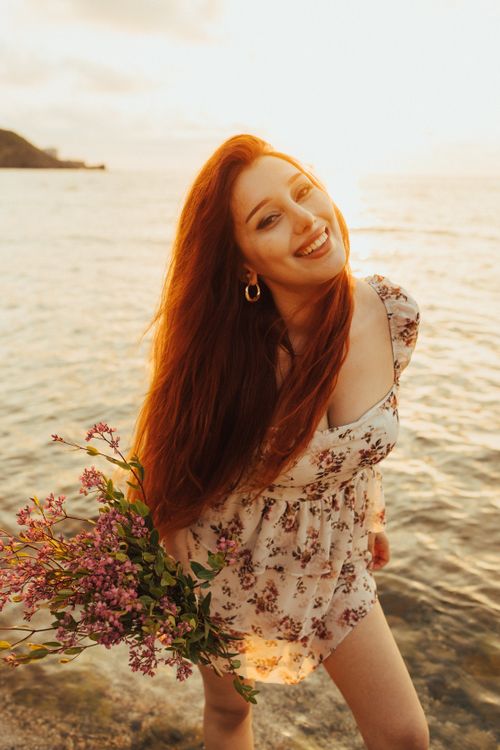 Free A cheerful woman with flowers by the sea during sunset in Türkiye, embodying relaxation and joy. Stock Photo