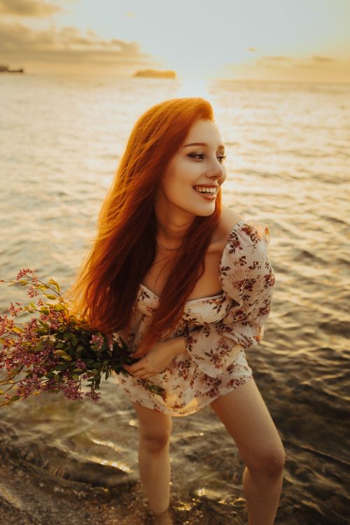 Free Joyful woman with red hair holding flowers by the sea during golden sunset in Türkiye. Stock Photo