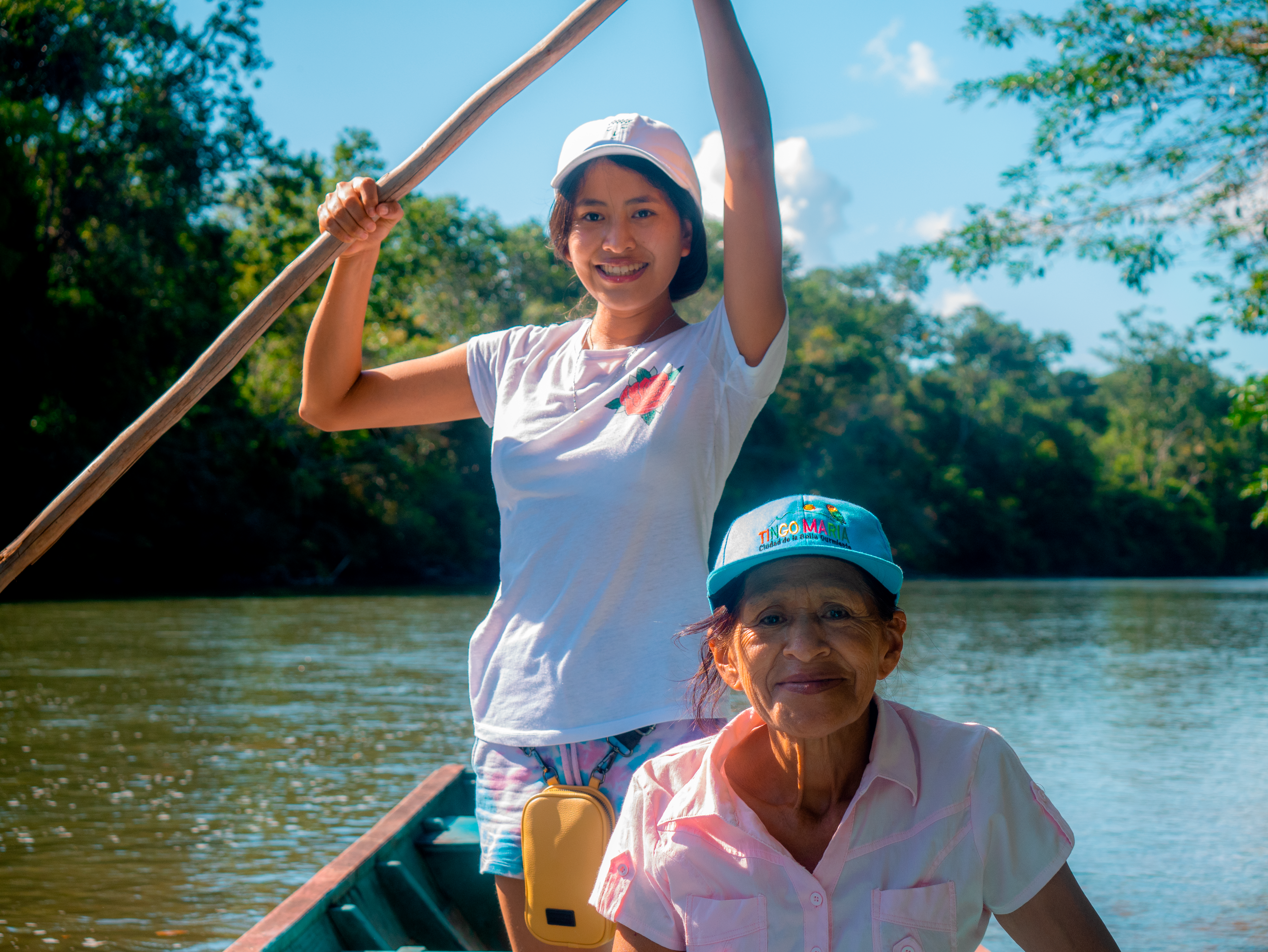 Two women enjoy a sunny day paddling a boat on a peaceful river surrounded by lush green trees.