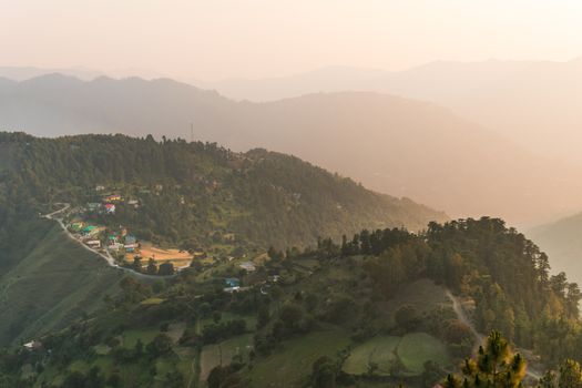 An aerial view of a picturesque mountain village surrounded by lush forest and rolling hills under a hazy sky.