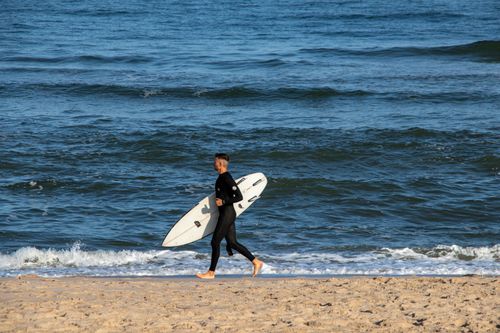 Free Man in wetsuit running on sandy beach carrying a surfboard by the ocean. Stock Photo