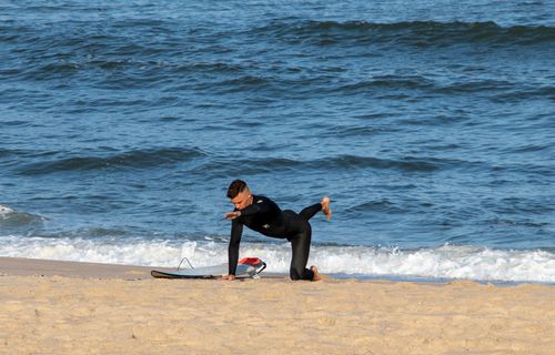 Free A man practicing yoga stretches on a sandy beach in Sylt with a surfboard nearby, under a clear sky. Stock Photo
