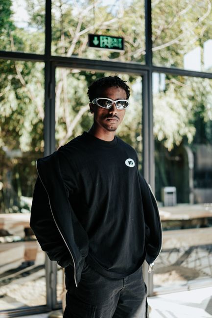 Fashionable man in black outfit and sunglasses posing indoors with natural light and greenery.