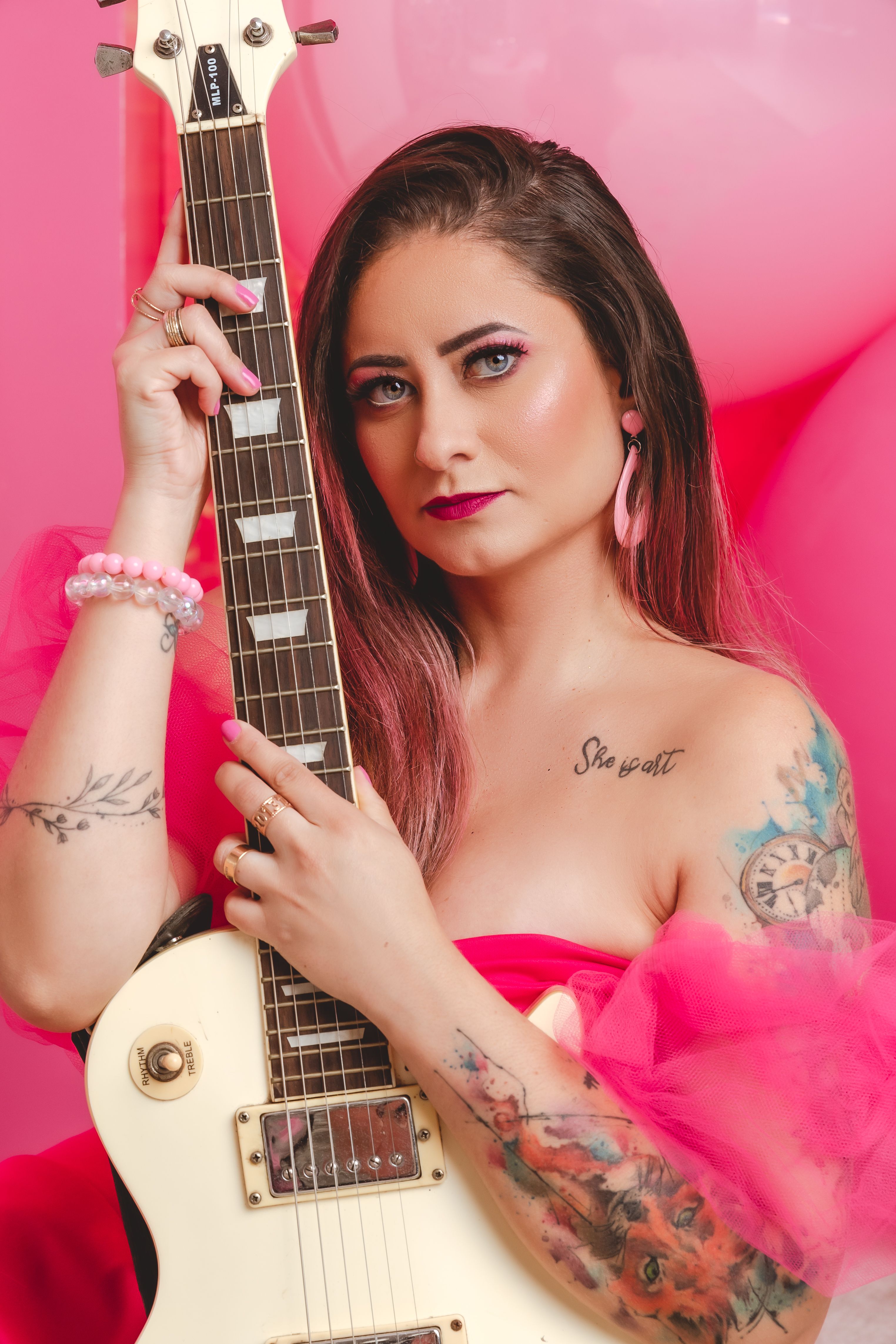 Portrait of a woman with tattoos holding an electric guitar against a pink backdrop.