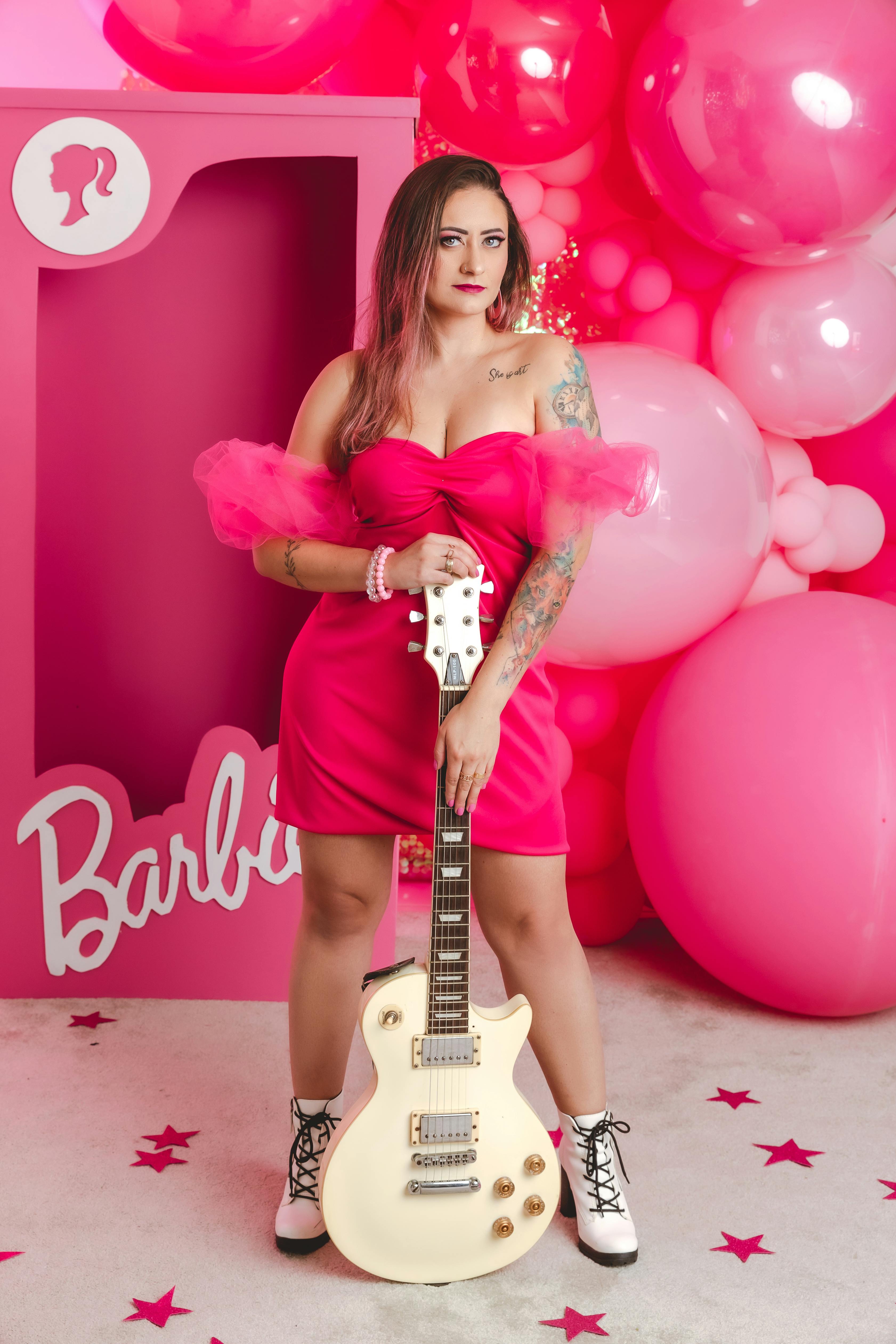 young woman with tattoos holding an electric guitar and standing on the background of pink balloons and a barbie box