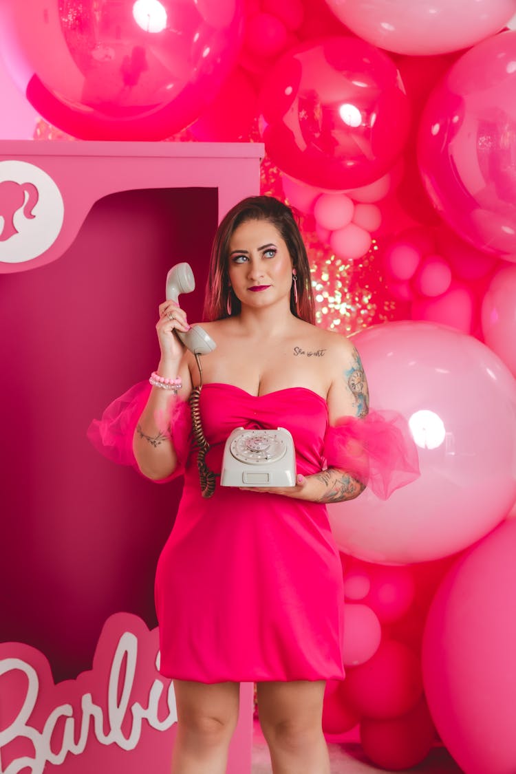 Young Woman In A Pink Dress Standing On The Background Of Pink Balloons And A Barbie Box