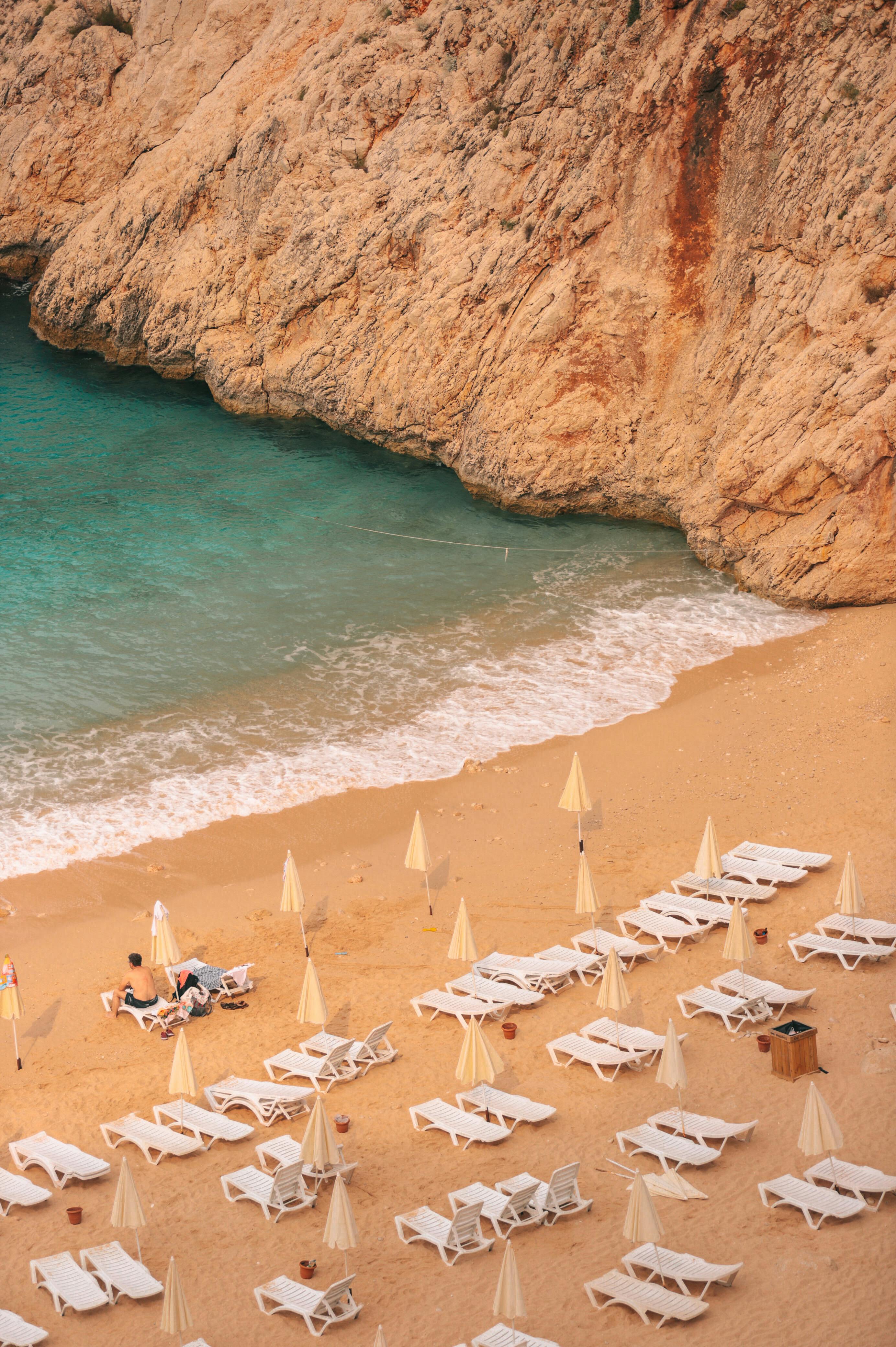 Aerial view of a tranquil beach with sunbeds, umbrellas, and calm turquoise waters meeting rocky cliffs.