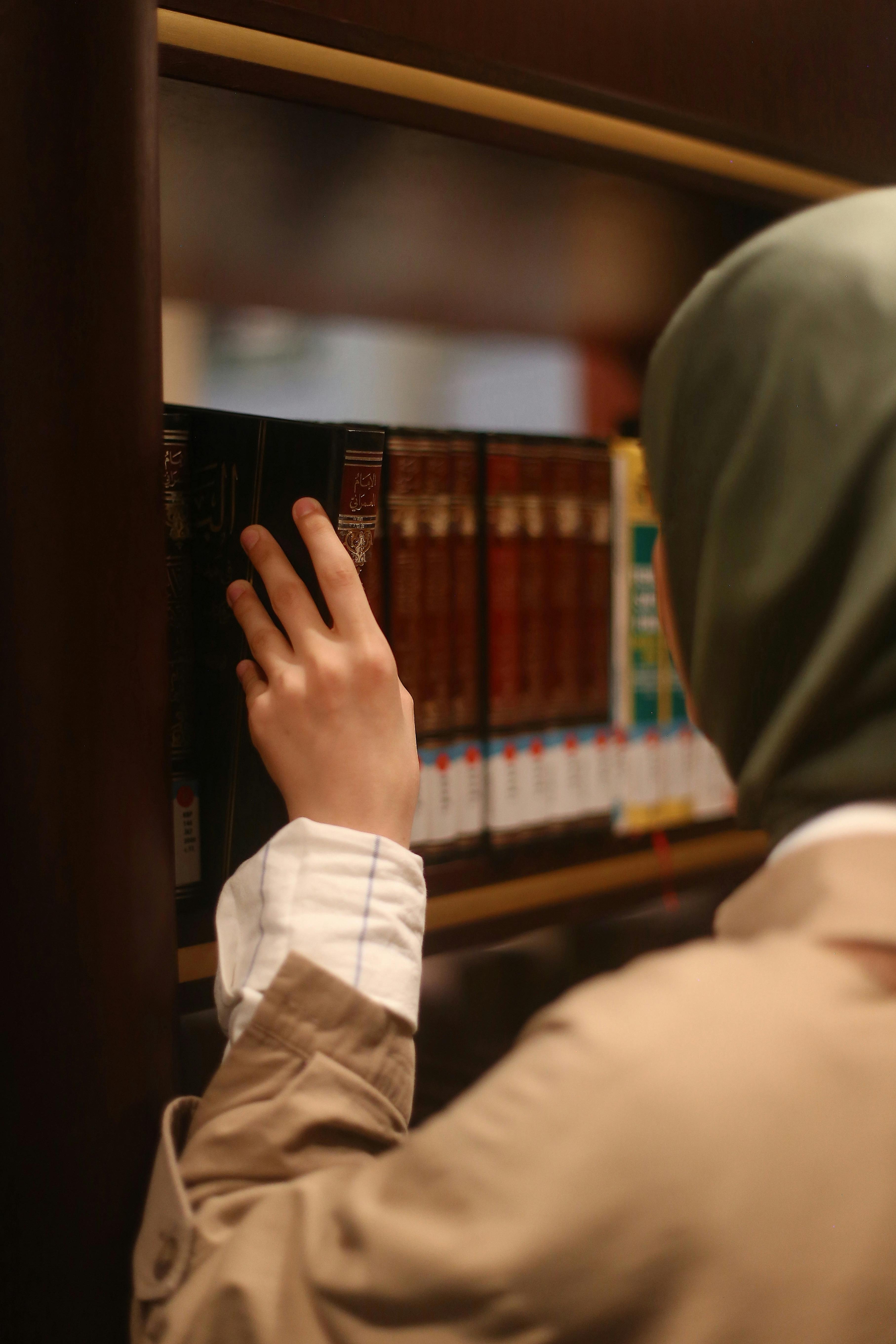 A woman reaches for a book on a library shelf, surrounded by volumes.
